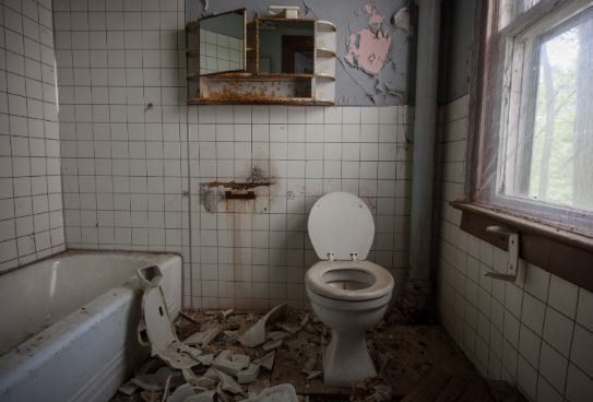 Dilapidated bathroom with broken tiles and rusty fittings, and messy floor.