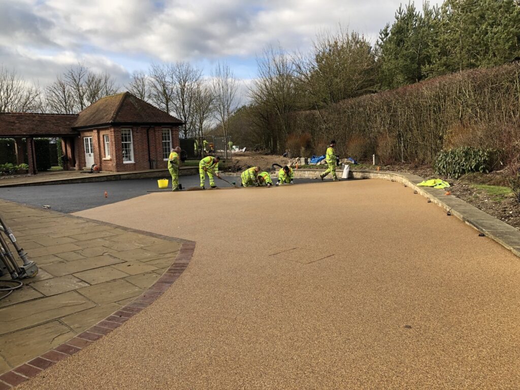 Workers in high-visibility gear laying a resin driveway near a brick building and garden, under cloudy skies.