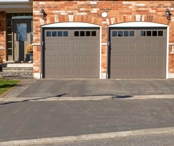 An asphalt driveway with two garage doors on a detached house made with orange bricks.